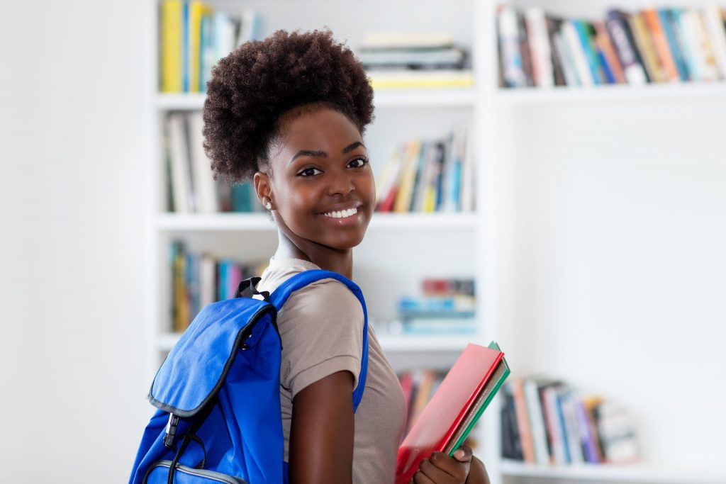 Uma estudante sorridente de pós-graduação, com uma mochila azul nas costas, segurando dois cadernos na mão.