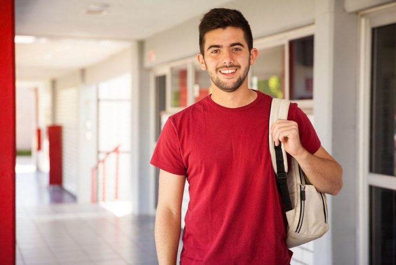 Um estudante pós-graduando de camisa vermelha carregando uma mochila e de pé em um corredor da universidade.
