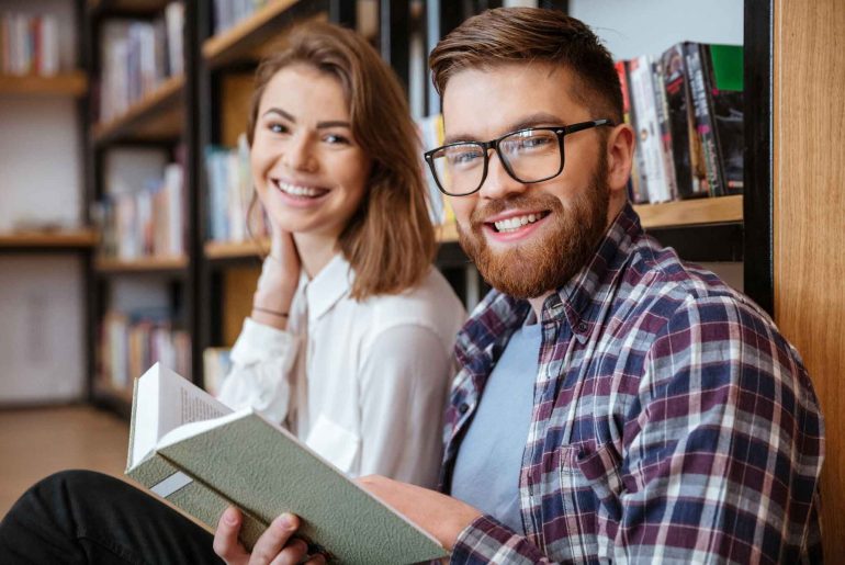 Dois estudantes pós-graduandos estudando juntos sentados no chão da biblioteca da universidade/faculdade, com um livro aberto em mãos.