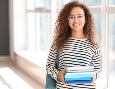 Uma estudante pós-graduanda sorridente, com uma mochila nas costas e segurando alguns livros.
