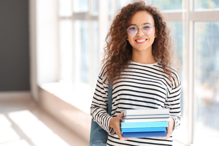 Uma estudante pós-graduanda sorridente, com uma mochila nas costas e segurando alguns livros.